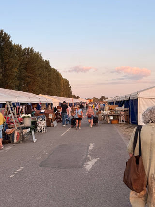 Shoppers pulling carts walk down a road between two long rows of white marquees at an outdoor market under a pink-tinged sky.