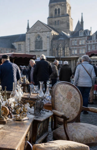A vintage-style upholstered chair in the foreground at an outdoor flea market, with antique tables, glassware, and people walking in the background near historic stone buildings.