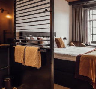 A modern bedroom interior with a dark wood slatted screen and towel bar holding two folded tan towels, with a bed and window in the background.