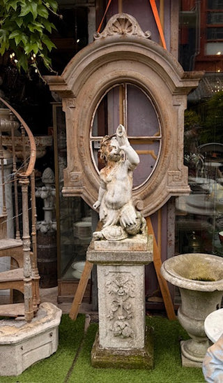 A classical-style stone statue of a nude female figure seated on a pedestal, displayed outdoors in front of a large decorative wooden arch and shop windows.