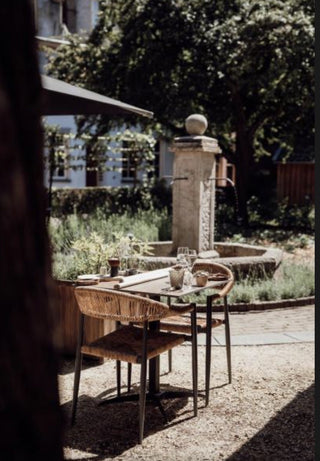 Outdoor patio dining setup with woven wicker chairs and a small table in a sunlit garden courtyard with a stone fountain and trees in the background.