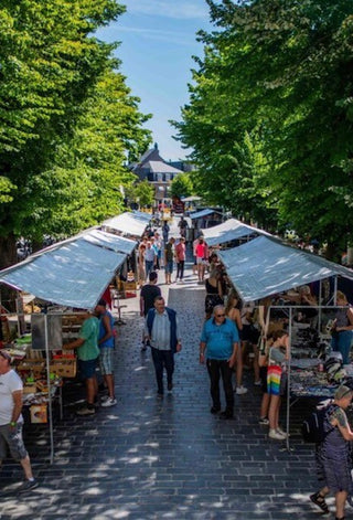A bustling outdoor market scene with rows of white canopy stalls lining a paved pathway between tall green trees, with people walking and shopping under a bright blue sky.