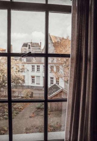 A view from inside a room looking out through a black-framed multi-pane window at an outdoor courtyard and historic white buildings, with a brown curtain on the right side.
