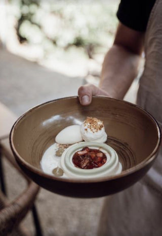 A person holding a rustic brown ceramic bowl containing a dessert with white cream, a meringue-like dollop, and a small red berry compote, photographed in a softly blurred real environment.