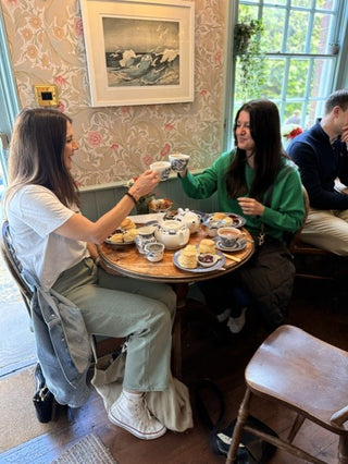Two women sitting at a wooden table in a cozy cafe setting, sharing tea and pastries. The room features vintage floral wallpaper, a framed landscape painting on the wall, and a large window in the background.