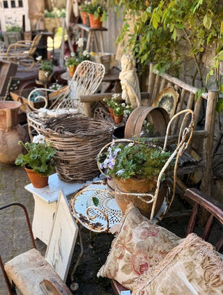 A rustic outdoor display of vintage furniture and home decor, featuring a large woven wicker basket, distressed metal chairs, potted plants, patterned cushions, and antique wooden pieces.