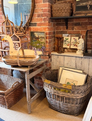 A rustic wicker basket filled with framed prints sits on the floor in a vintage-style room with a brick wall, wooden table, and other decorative home items.