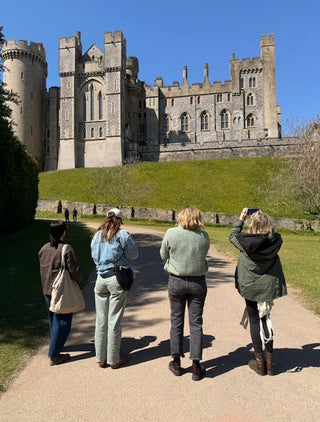 Four people standing on a path in front of a large stone castle on a grassy hill under a clear blue sky.