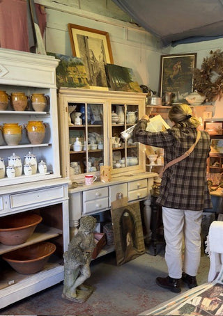 A woman in a plaid shirt and white pants browses an antique cabinet filled with ceramic dishes in a cluttered vintage furniture shop.