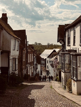 A scenic photograph of a narrow cobblestone street lined with old white and brown houses under a cloudy sky, with a few small people walking in the distance.