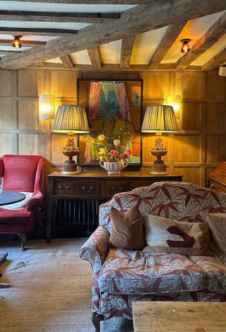 A cozy, rustic living room featuring a patterned armchair in the foreground, a red armchair on the left, and a dark wood console table with two lit table lamps and a colorful painting under exposed wooden ceiling beams.