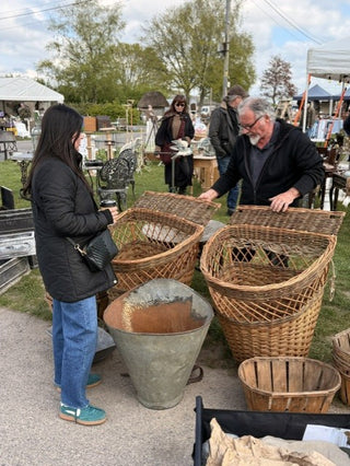Two people standing outdoors at what appears to be a flea market or antique show, examining large woven wicker baskets. One person wears a black puffer jacket and blue jeans, and the other wears a dark jacket.