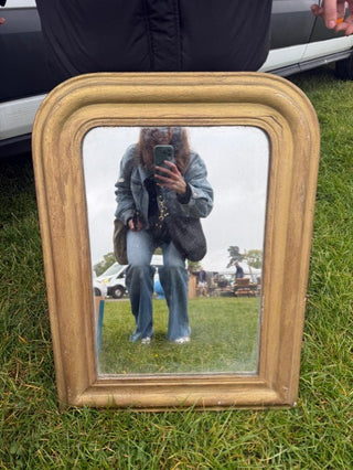 A rectangular vintage-style mirror with a distressed gold frame, resting on green grass. The mirror reflects a person taking a selfie outdoors.