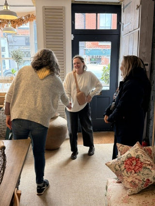 Three women standing and interacting inside a home interior with a black front door, windows, a rug, and floral pillows visible.