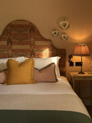 A cozy bedroom scene featuring a bed with white linens, a mustard yellow pillow, and a patterned headboard under warm bedside lighting.