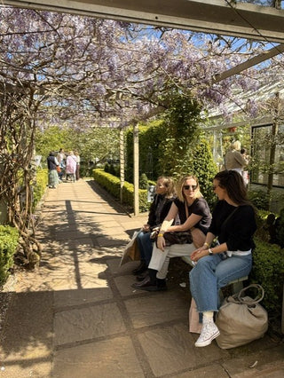 Three women sitting on a bench in a sunlit garden conservatory walkway under a wisteria canopy, with other visitors and greenery in the background.