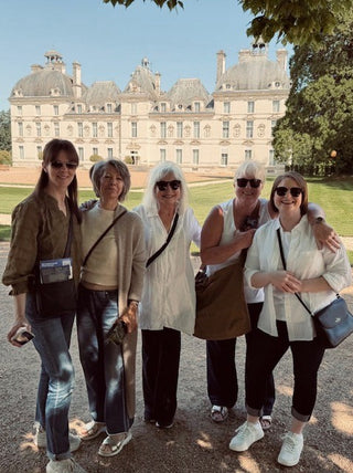 Five women posing together outdoors in front of a large historic chateau, wearing casual summer outfits and carrying crossbody bags.