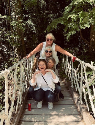 Four women posing together on a rustic wooden bridge with white railings in a lush green forest.