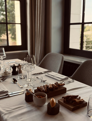 A dining table set with a white tablecloth, wine glasses, cutlery, and small wooden boards holding food, photographed in a softly lit indoor dining or restaurant setting with windows and chairs in the background.