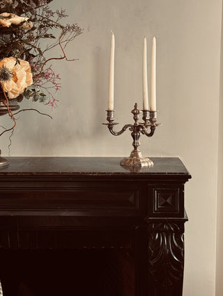 A three-arm candle holder with a dark metallic finish sits on a dark wooden mantelpiece, holding three white taper candles. To the left, a dried floral arrangement is partially visible against a neutral gray wall.