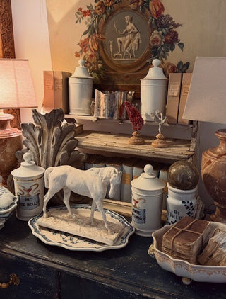 A styled vintage interior scene with multiple ceramic canisters, decorative boxes, a white horse figurine on a plate, books, lamps, and a floral wall hanging.