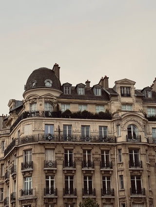 A classic Parisian-style apartment building facade with ornate stone detailing, black wrought-iron balconies, multiple windows, and a dark dome roof under a pale sky.