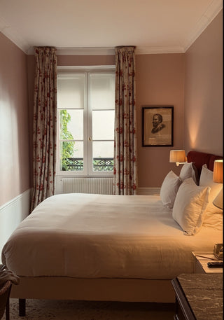 A neatly made bed with white linens and multiple pillows in a softly lit bedroom with dusty pink walls, patterned curtains, a window, and bedside lamps.