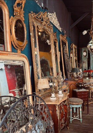 A collection of ornate vintage mirrors with gilded frames displayed on a dark teal wall in an antique shop. The scene includes a carved wooden console table with a marble top, a light blue stool, and other antique furniture pieces.