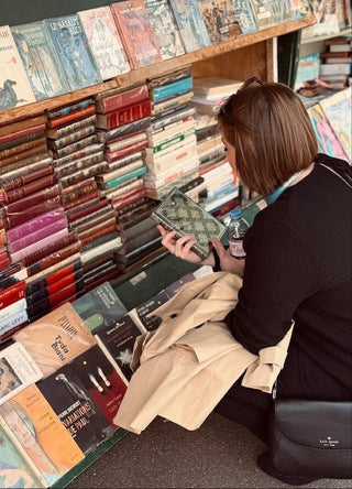 A woman with short brown hair browses a bookshelf in a bookstore, holding a book and wearing a black top, beige coat, and a black Kappa shoulder bag.