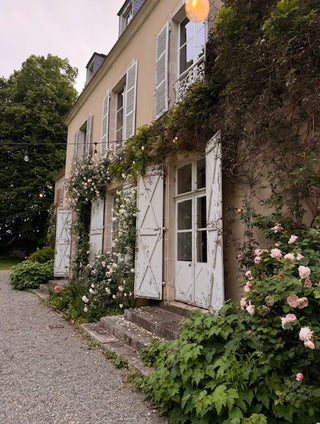 A charming beige stone building with white shutters, heavily adorned with climbing green vines and pink roses, set beside a gravel path.