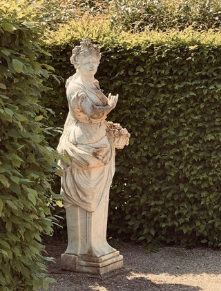 A classical-style stone garden statue of a draped female figure holding a bouquet, set outdoors against a dense green hedge and ivy-covered wall.