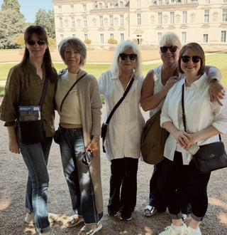 Five women posing together outdoors in front of a large classical building, each wearing sunglasses and carrying black crossbody bags.