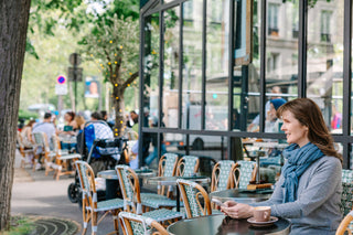 A woman sits at an outdoor cafe table next to a large glass window, wearing a gray top and a blue scarf. The cafe has patterned chairs and tables, with trees and other patrons visible in the background.