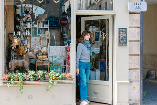 A woman wearing a light blue-gray sweater, blue jeans, and a blue scarf stands in the doorway of a quaint boutique shop. The shop window displays various home decor items, candles, and potted plants, while a street sign reading 'SAUF' is visible on the right.
