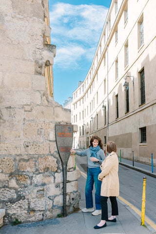 Two women standing on a city street beside a stone wall and informational sign, with historic buildings and a blue sky in the background.