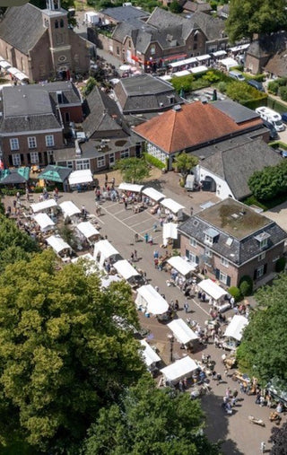 Aerial view of a small town square with rows of white market tents, people walking, and surrounding historic buildings with gray and terracotta roofs.