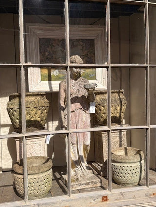A classical-style stone garden statue of a draped female figure stands behind a large wooden grid window, flanked by large decorative urns and two smaller planter baskets.