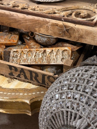 A close-up of ornate vintage-style carved wood pieces and decorative trims, including a carved block with figures, a wooden sign reading PARIS, and a gray woven textile with clear rhinestones in the foreground.
