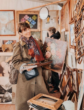 Woman holding a painting in an art studio with various artworks and tools around.