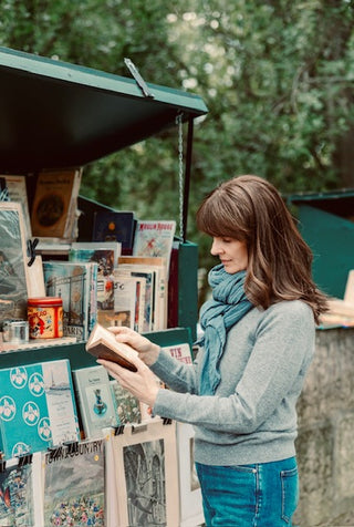 A woman with brown hair wearing a gray sweater and blue scarf reads at an outdoor book stall filled with books and small items.